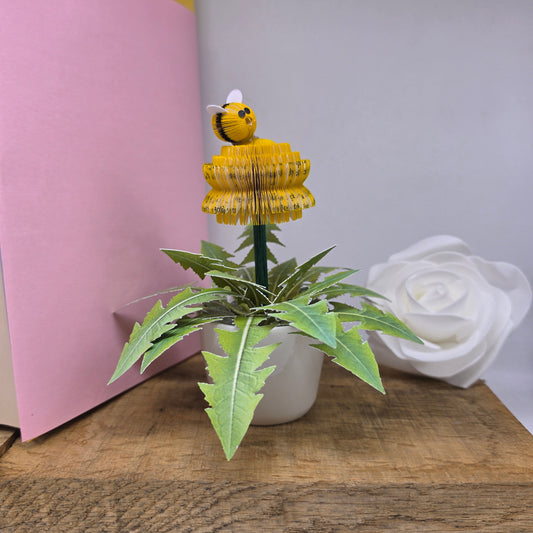 A handmade paper flower sculpture featuring a yellow striped bumblebee on a layered yellow flower stem, with green layered leaves below, displayed on a wooden surface next to a pink card and a white paper rose.